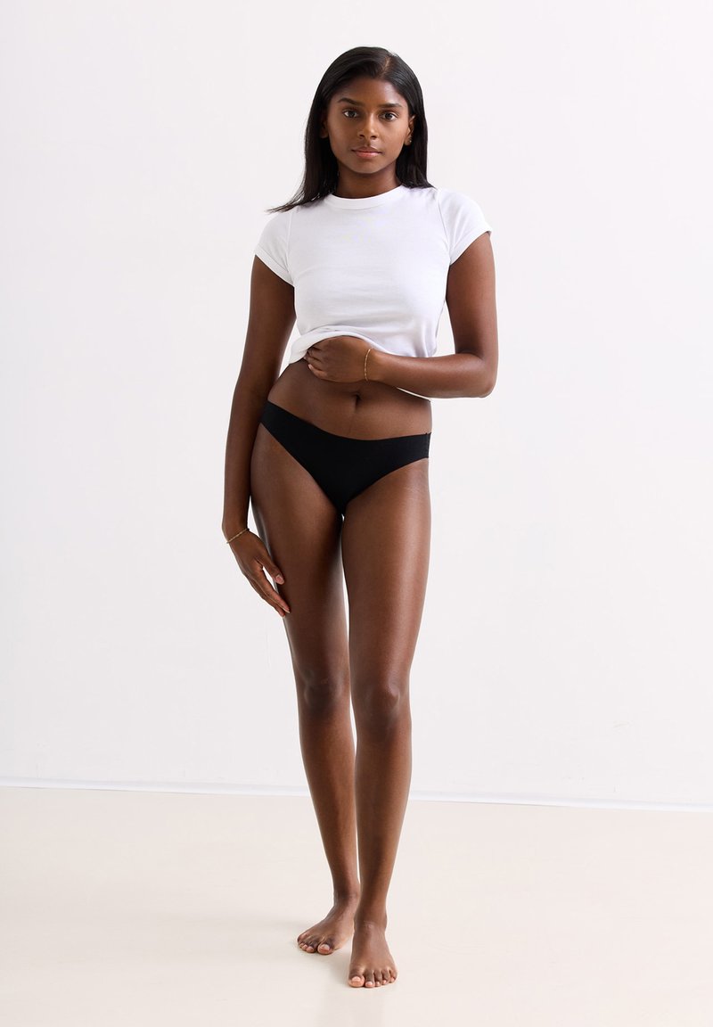Witte getailleerde t-shirt, zwarte lingerie slips. Model heeft steil donker haar en staat blootvoets met ontspannen armen aan haar zijde. Neutrale achtergrond.