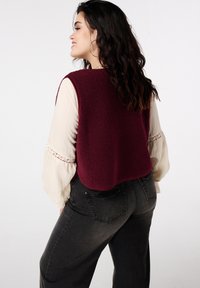 Veste sans manches en polaire bordeaux avec un col rond, portée sur un chemisier crème avec des manches texturées et des détails en dentelle, associée à un jean gris foncé.