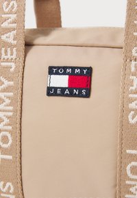 Geantă din nylon bej, cu un patch proeminent "TOMMY JEANS" în culorile navy, roșu și alb, având curele țesute care afișează numele mărcii în text bej.