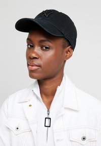 Gorra de béisbol negra con logo bordado, visera curva y correa ajustable en la parte trasera, llevada con una chaqueta blanca y una parte superior con cremallera de canalé.