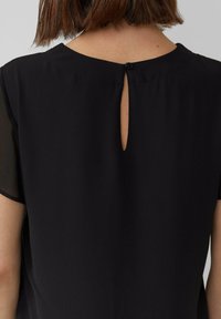 Vrouw draagt een zwarte blouse met korte mouwen en een sleutelgat aan de achterkant, gesloten met een enkele knoop bij de halslijn.