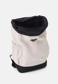 Sac à dos avec un corps en tissu beige doux et un sommet noir. Comprend une ouverture cintrée, une poche latérale et des bretelles noires. Surface texturée.