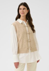 Žena v béžovej pletenej veste s mašľovými detailmi, oblečenej na bielu košeľu s dlhým rukávom, stojí pred jednofarebným svetlým pozadím.