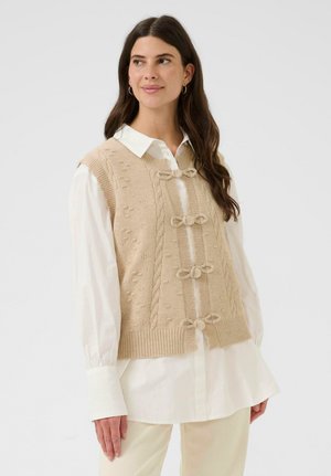 Eine Frau, die einen beigen Strickpullover mit Schleifen-Details über einem weißen Langarmshirt trägt, steht vor einem einfachen hellen Hintergrund.