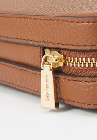 Esquina de una bolsa de cuero marrón texturizado con un tirador de cremallera dorado que lleva una etiqueta grabada con "MICHAEL KORS". Costuras simples a lo largo de los bordes.