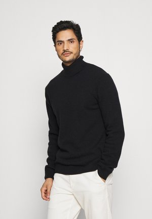 Suéter negro de cuello alto hecho de un tejido de punto texturizado, con un diseño ajustado y puños acanalados, combinado con pantalones de color claro.