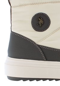 Beige syntetiskt överdel med mörkgrå accenter, med texturerade material och en cirkulär logotyp-patch. Tjock vit mellansula med greppvänlig yttersula.