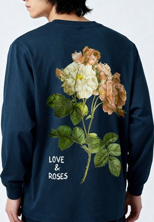 Mann trägt ein marineblaues Langarmshirt mit großem Aufdruck eines Straußes aus rosa und weißen Rosen und dem Text „LOVE & ROSES“ auf dem Rücken.