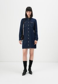 Femme debout portant une robe en denim bleu foncé à boutons, des chaussettes noires mi-mollet, des escarpins à lacets noirs et un petit sac à bandoulière noir.