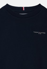 Tums zila crewneck sudraba krekliņa izgatavots no kokvilnas, ar ribotu apkakli un nelielu baltu logotipu uz krūtīm, ar sarkaniem un baltajiem akcentējošiem svītrojumiem.