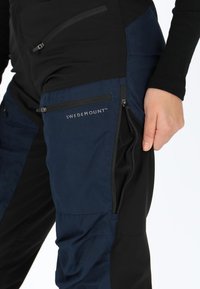 Marineblauwe en zwarte broek met een zachte textuur, uitgerust met meerdere ritszakken en het "SWEDEMOUNT"-logo op de dij.