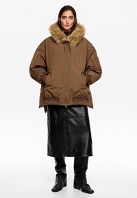 Chaqueta marrón con aislamiento y capucha forrada de piel, cremallera delantera, puños ajustados y bolsillos laterales, combinada con una falda de cuero negra y botas.