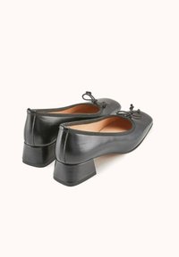 Des chaussures à talon bloc en cuir noir présentent un bout arrondi et un détail de nœud décoratif à l'avant. Texture lisse avec un subtil éclat.