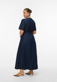 Mujer con cabello castaño recogido en un moño, vestida con un vestido largo de mezclilla azul oscuro y zapatos slingback de tacón bajo color beige, de pie contra un fondo blanco.