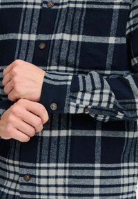 Niebiesko-biała koszula flanelowa w kratę z guzikami przy mankietach i kieszenią na piersi, charakteryzująca się teksturowanym materiałem i kontrastowymi wzorami kratki.