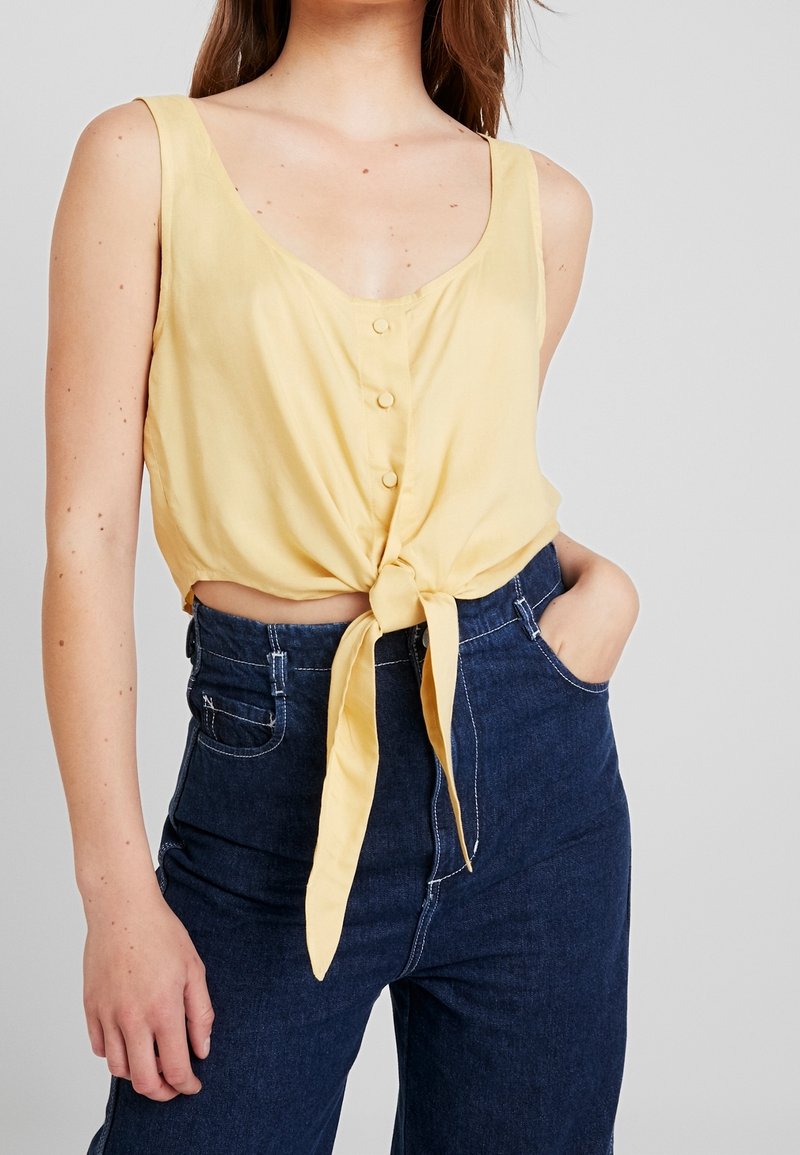 Gelbes ärmelloses Oberteil mit V-Ausschnitt und Knopfdetail, mit einer vorderen Schleife. Kombiniert mit hoch taillierten dunkelblauen Jeans.
