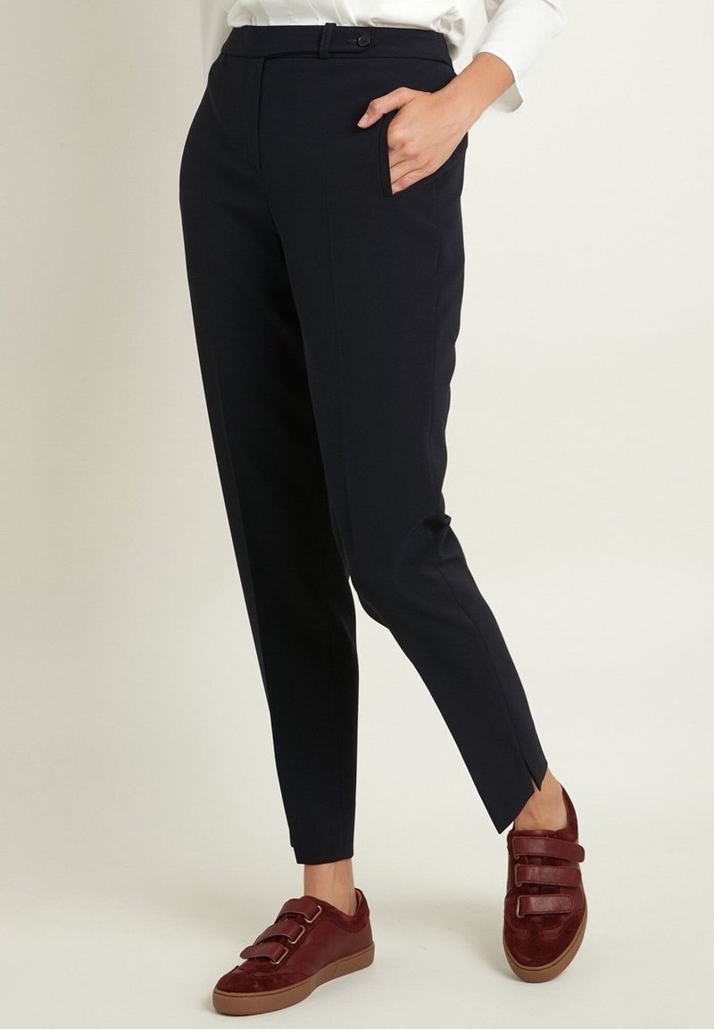 Personne portant un pantalon noir ajusté avec un bouton frontal et des poches, associé à des baskets bordeaux à scratch.