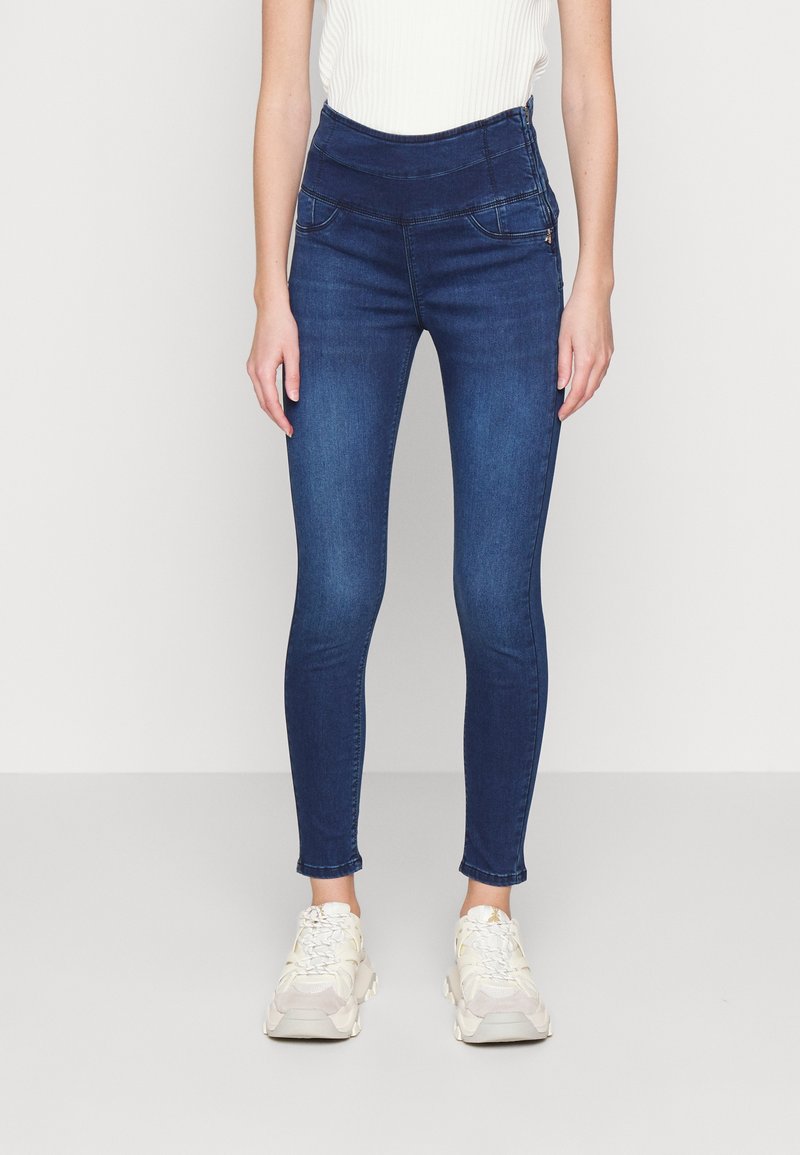 Slim-fit mørkeblå jeans med høj talje båret af en person, der står oprejst, kombineret med hvide chunky sneakers og en hvid ærmeløs top.
