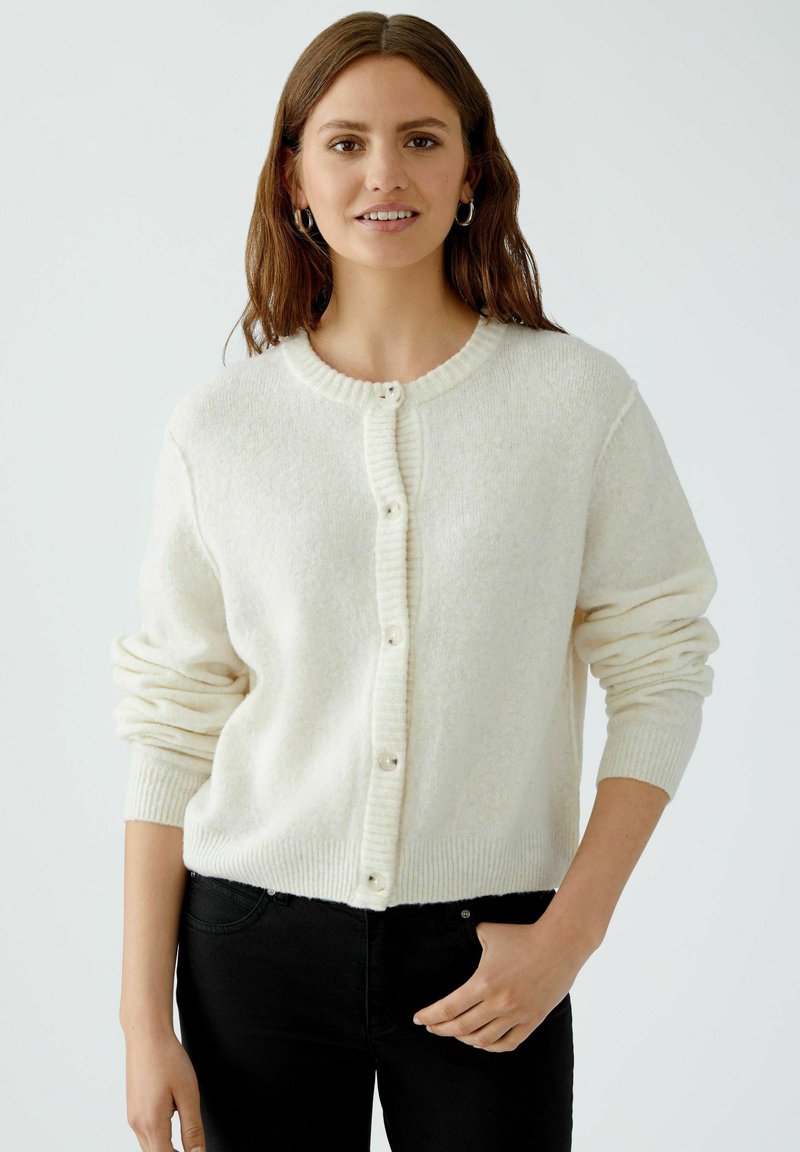 Helle beige Strickjacke aus weichem Material. Verfügt über einen runden Ausschnitt, gerippte Bündchen und eine Knopfleiste auf der Vorderseite.