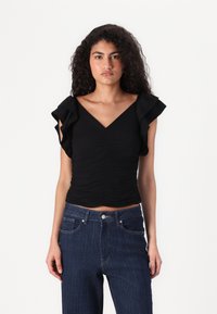 Svart topp med V-hals og volangermslommer, med rynkedetalj. Kombinert med mørke denimjeans med rette ben.