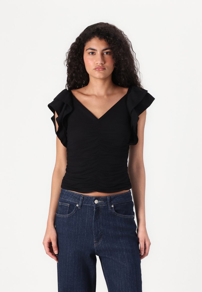 Svart topp med V-hals og volangermslommer, med rynkedetalj. Kombinert med mørke denimjeans med rette ben.