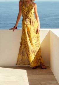 Robe longue jaune à imprimés floraux, col halter, taille nouée, tissu fluide et sandales. En arrière-plan, un océan bleu et un ciel dégagé.