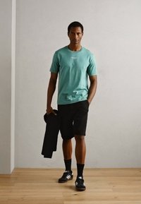 Menta zöld rövid ujjú póló, fekete rövidnadrág és fekete sportcipő. Fekete dzsekit tart, világos fa padlón áll egy szürke fal előtt.