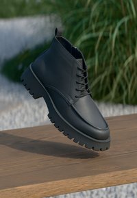 Bottine noire en cuir à lacets avec une semelle épaisse à crampons, flottant au-dessus d'une surface en bois avec des plantes vertes en arrière-plan.