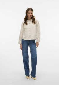 Beige kort jacka med knäppning framtill, två framfickor och krage, i kombination med blå raka jeans och krämfärgade klackar.