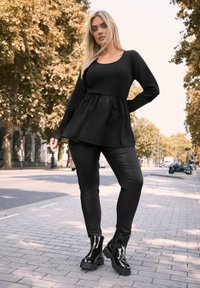 Musta peplum-paita pitkähihaisilla, yhdistettyinä kiiltäviin mustiin skinny-housuihin ja kiiltäviin nilkkasaappaisiin. Kaupunkimaisema, taustalla puita.