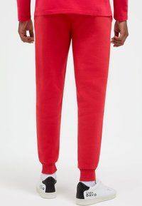 Mann, der rote Jogginghosen und weiße Turnschuhe mit schwarzen Fersenakzenten trägt, steht von hinten vor einem schlichten weißen Hintergrund.