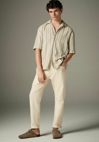 Camisa de manga corta a rayas en beige claro y blanco, combinada con pantalones beige pálido y zapatos de lona marrones, con un ajuste casual.