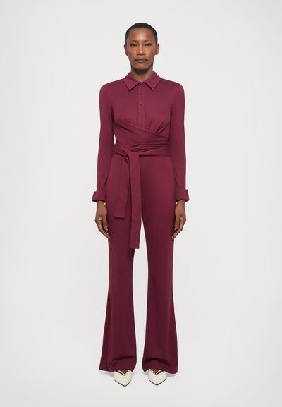 Salopette bordeaux con maniche lunghe, colletto e design avvolgente. Presenta una cintura in vita e pantaloni a zampa. Abbinata a scarpe bianche a punta.