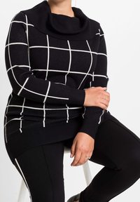 Pull en tricot noir avec col bénitier, présentant un motif à carreaux blancs. Porté avec des leggings noirs, offrant un contraste de texture et de design.