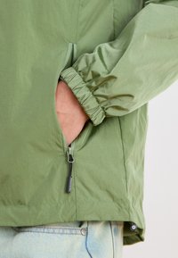 Green lightweight jacket with elastic cuffs, zippered pocket, and smooth texture. The design features a simple, functional style.