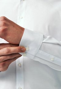 Chemise blanche avec une texture lisse, un col classique et des poignets à boutons. La main boutonne le poignet, montrant le détail du tissu.