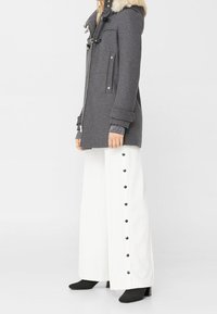 Manteau en laine gris avec capuche en fausse fourrure, fermetures éclair et à bouton, deux poches avant. Pantalons larges blancs avec des détails de boutons sur le côté, bottes noires à cheville.