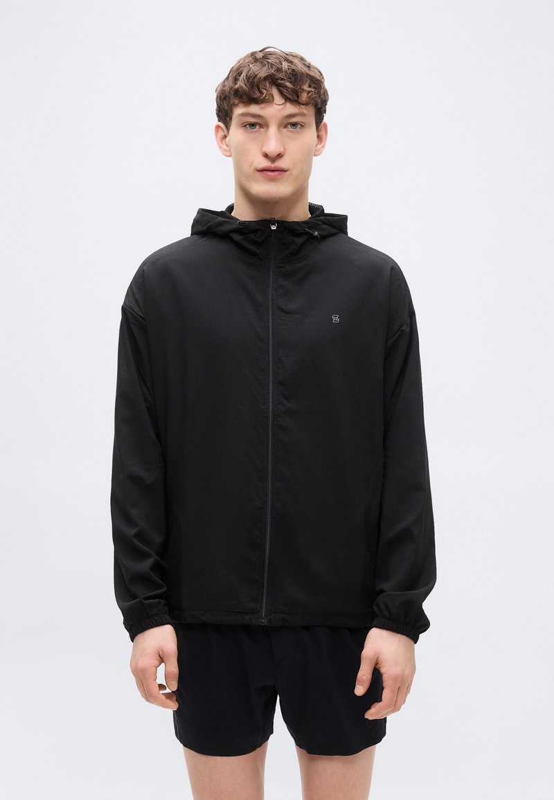 Μαύρο αδιάβροχο μπουφάν με φερμουάρ μπροστά, κουκούλα και ελαστικά ρεβέρ. Απλός σχεδιασμός με μικρό λογότυπο στο στήθος. Ελαφρύ υλικό.