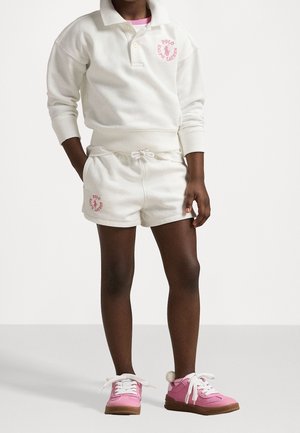 Dziecko ubrane w białą bluzę Polo Ralph Lauren i szorty z różowym logo oraz różowe trampki ze białymi sznurowadłami i pomponami.