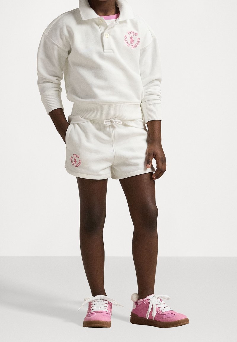Enfant portant un sweat-shirt Polo Ralph Lauren blanc et un short avec un logo rose, ainsi que des baskets roses avec des lacets blancs et des pompons.