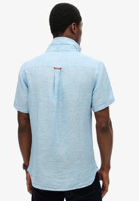 Hombre de espaldas, vistiendo una camisa de manga corta azul claro con una pequeña etiqueta naranja en la parte superior de la espalda, combinada con pantalones oscuros.