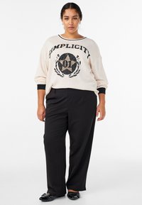 Krēmkrāsas džemperis ar melnu tekstu "Vienkāršība" un zvaigznes grafiku, kombinēts ar melnām biksēm un melniem apaviem ar dekoratīvām detaļām.