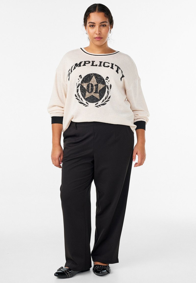 Krēmkrāsas džemperis ar melnu tekstu "Vienkāršība" un zvaigznes grafiku, kombinēts ar melnām biksēm un melniem apaviem ar dekoratīvām detaļām.