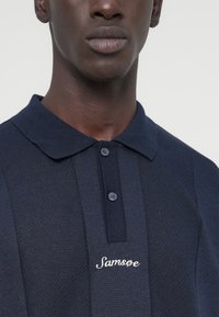 Polo bleu marine en tissu texturé, avec deux boutons et un logo "Samsoe" brodé sur le côté gauche de la poitrine. Col classique.