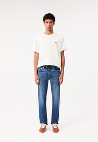T-shirt bianco in cotone con un piccolo logo verde, jeans in denim blu, cintura nera e sneakers arancioni con lacci bianchi.