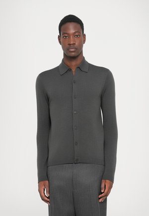 Hombre con cabello corto, vistiendo un cárdigan de botón gris oscuro y pantalones gris a juego, de pie contra un fondo claro y liso.