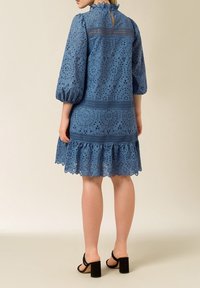Vestido de broderie anglaise azul con mangas abullonadas, dobladillo con volantito y detalle de ojal en la espalda. Textura suave y patrones florales intrincados visibles.