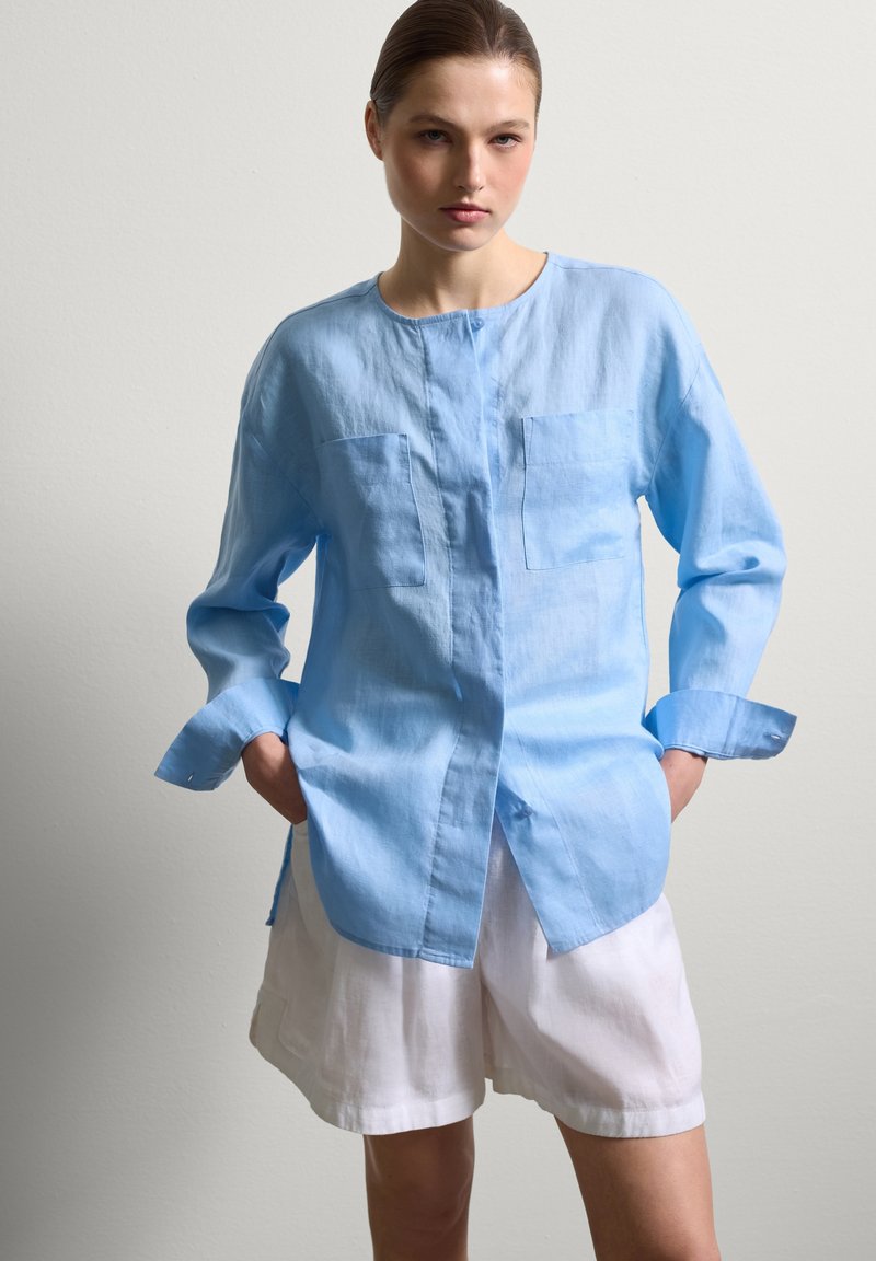 Helles blaues Leinenhemd mit langen Ärmeln, Stehkragen und zwei Brusttaschen. Kombiniert mit weißen Leinen-Shorts, die eine lässige Passform zeigen.