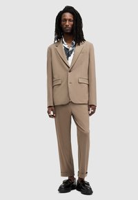 Traje beige con americana estructurada y pantalones a medida. Zapatos negros brillantes, camisa de patrón debajo y puños enrollados en los pantalones.