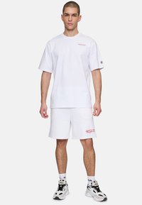 T-shirt en coton blanc à coupe décontractée, texte rouge sur la poitrine, associé à un short blanc avec le logo "R.S.V.P." rouge, complété par des baskets noir et blanc.